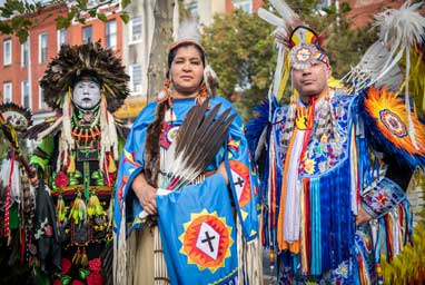  A woman and two men in Indigenous festival attire stand in front of the Baltimore American Indian Center.